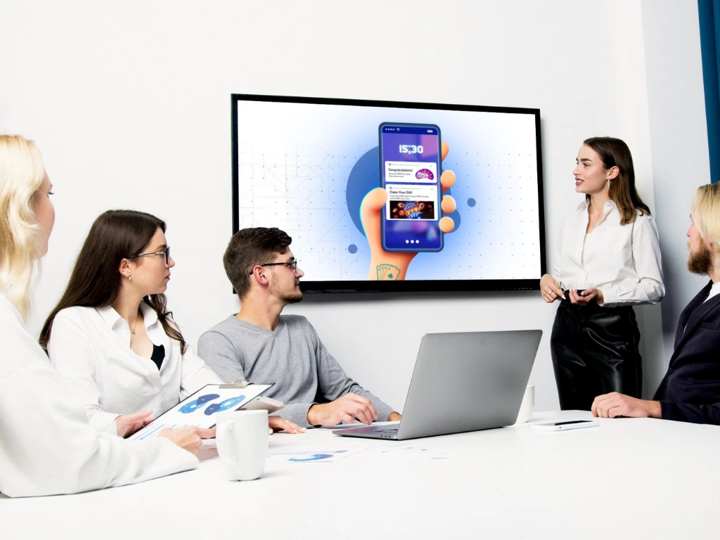 Four people sit at a conference table with laptops and papers, watching Tim Bilyeu present a mobile app interface displayed on a large screen at the front of the room. 